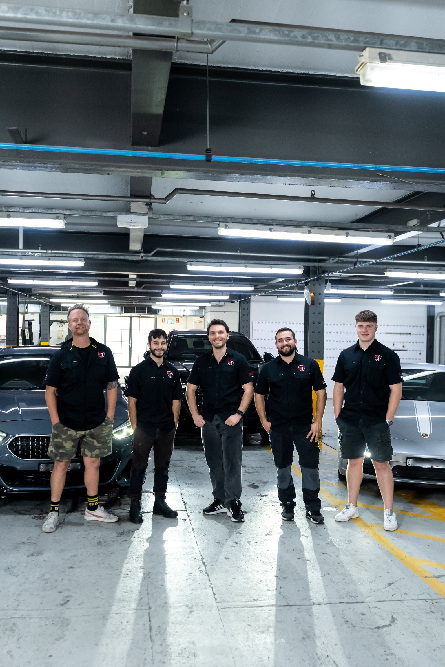 Five GenTek car protection specialists standing casually with their hands in their pockets beside luxury vehicles.