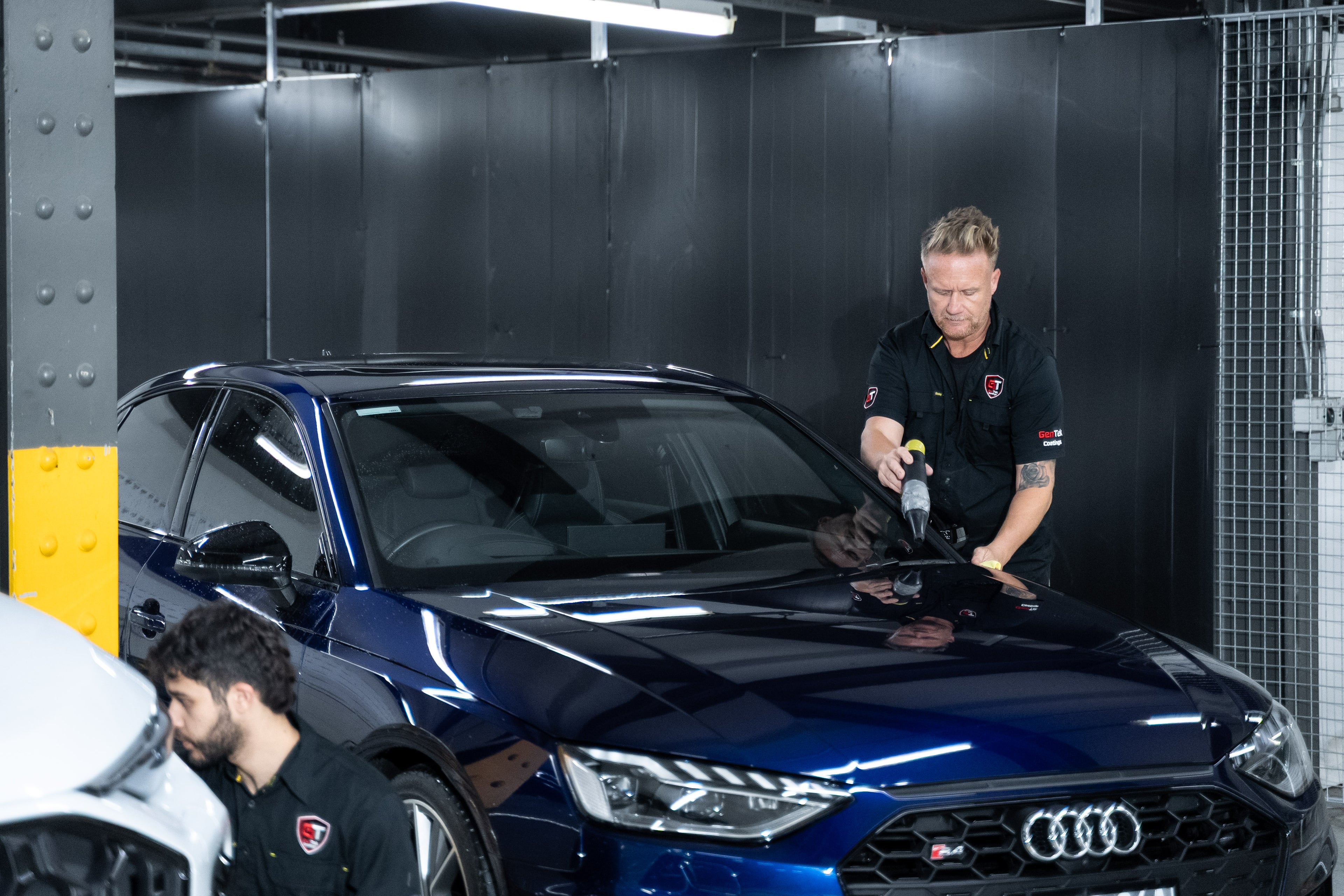 A dark metallic blue Audi car in a professional garage setting, with two technicians working on it. One technician is polishing the car's hood with a microfiber cloth while the other is visible in the foreground.